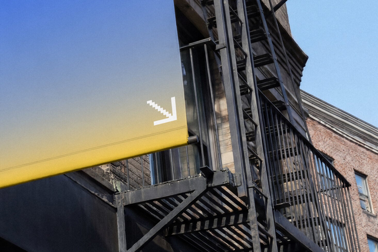Bright yellow storefront awning with minimalist arrow logo mounted on industrial metal fire escape against brick building