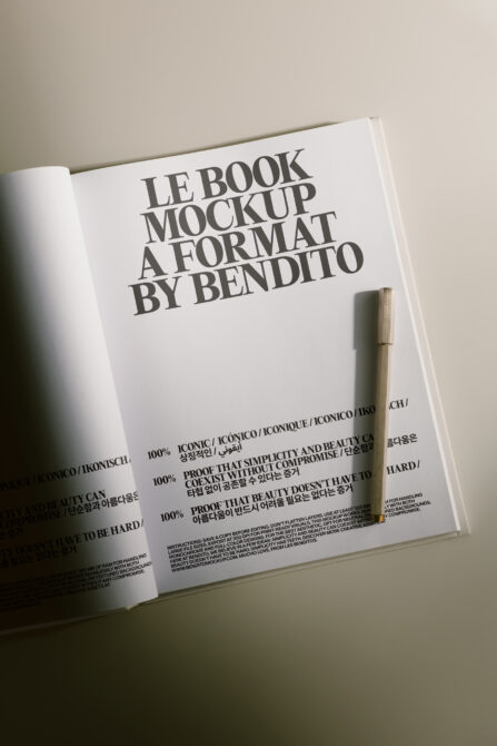 Open magazine spread showing Le Book mockup with bold typography, multilingual text, plus wooden pen on neutral background