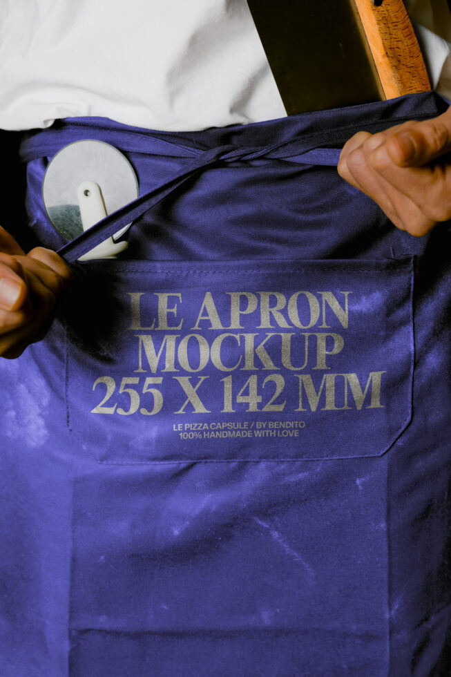 Hands opening deep blue apron with gold text, wooden spoon peeking out from pocket