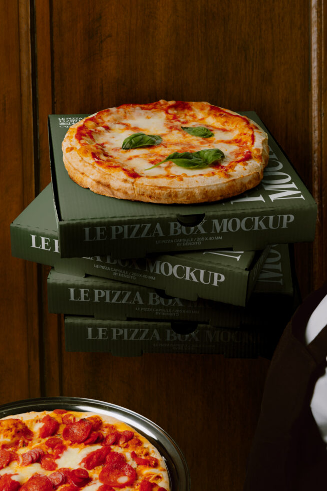 Stacked green pizza boxes with fresh margherita on top, pepperoni pie below on wooden backdrop