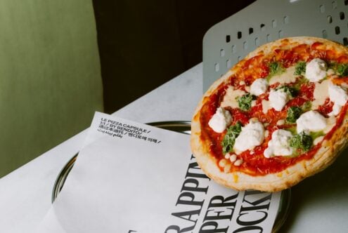 Fresh pizza on multilingual packaging with green background, perforated metal basket visible