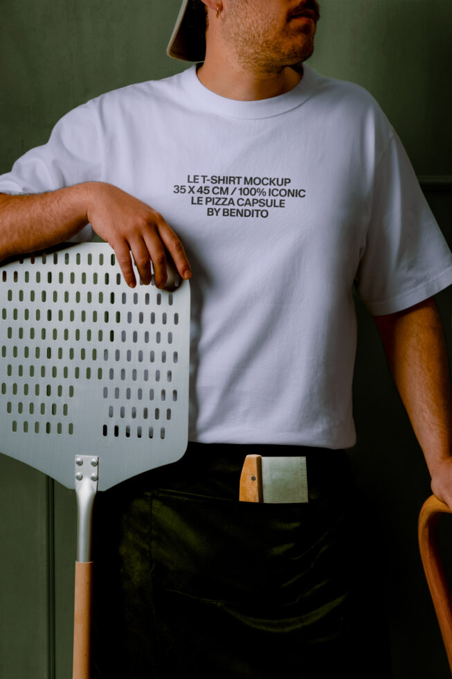Guy in white tee holding pizza peel against sage green wall, mockup ready to customize