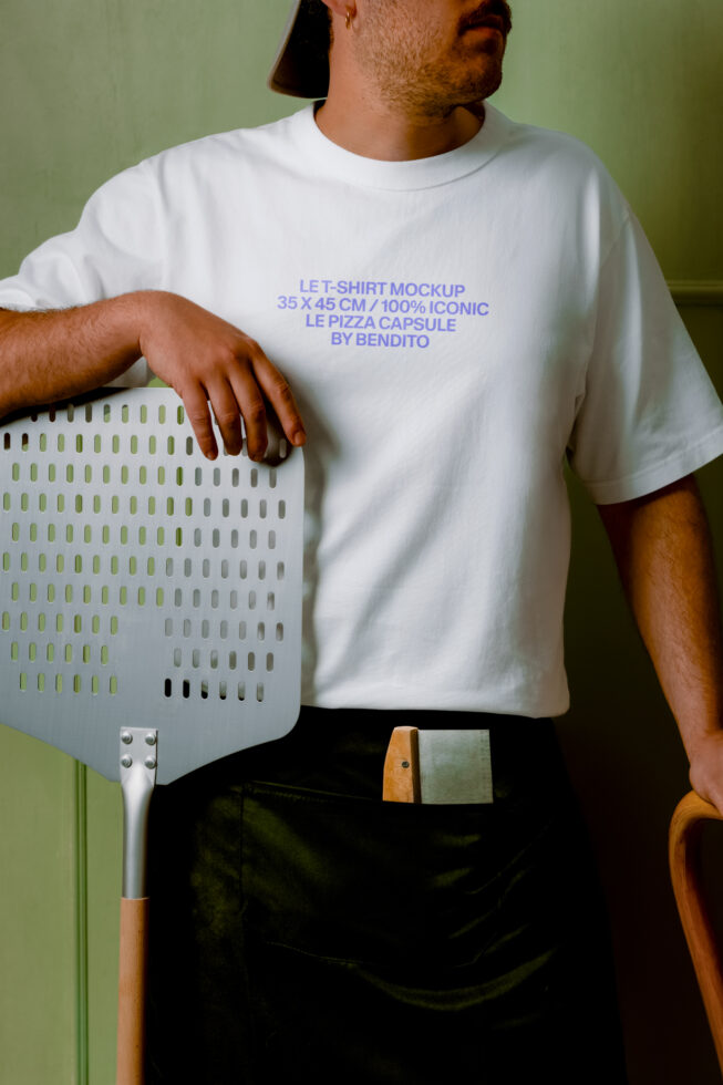 Guy in white tee holding pizza peel against sage green wall, periwinkle text mockup