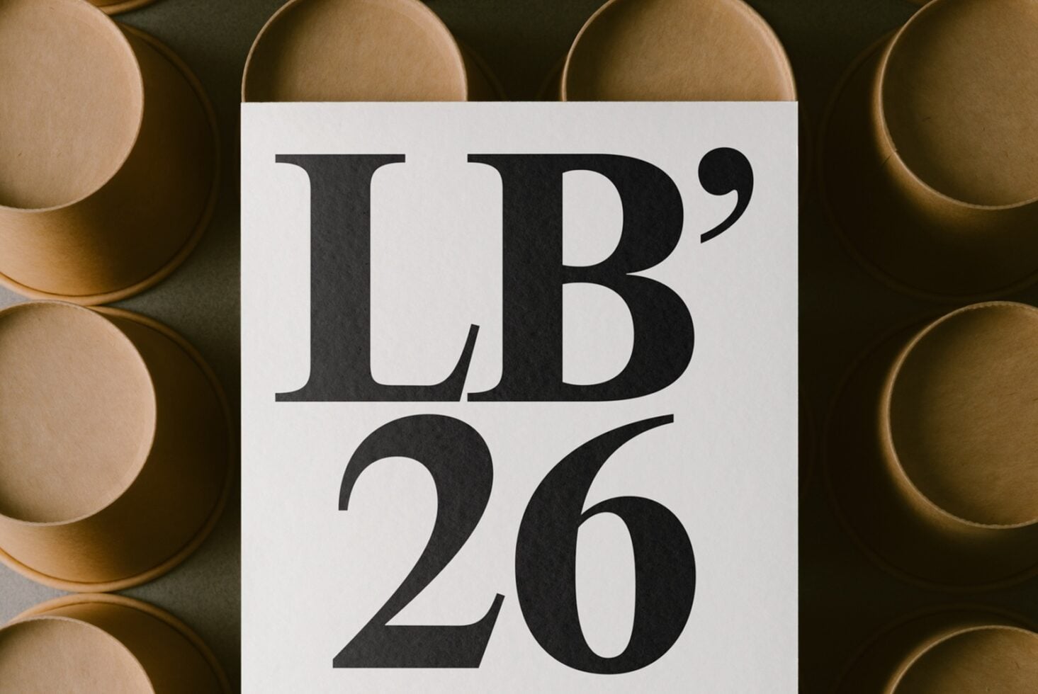 White card with bold serif typography displaying "LB'26" surrounded by stacked brass bowls on dark background