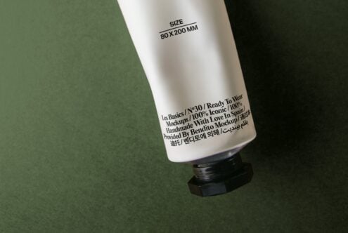 White cosmetic tube with black cap on sage green background showing multilingual label