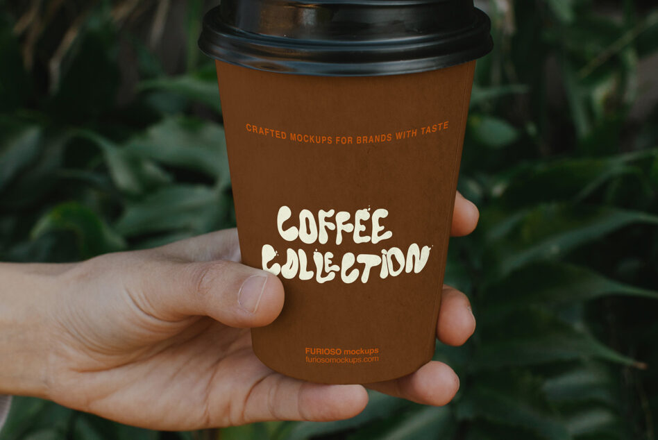 Hand holding brown paper coffee cup with cream text logo against blurred green foliage