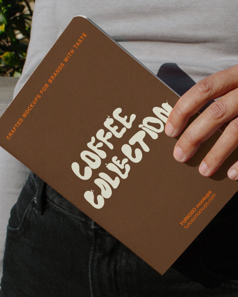Hand holding brown coffee table book mockup with cream lettering in sunlight