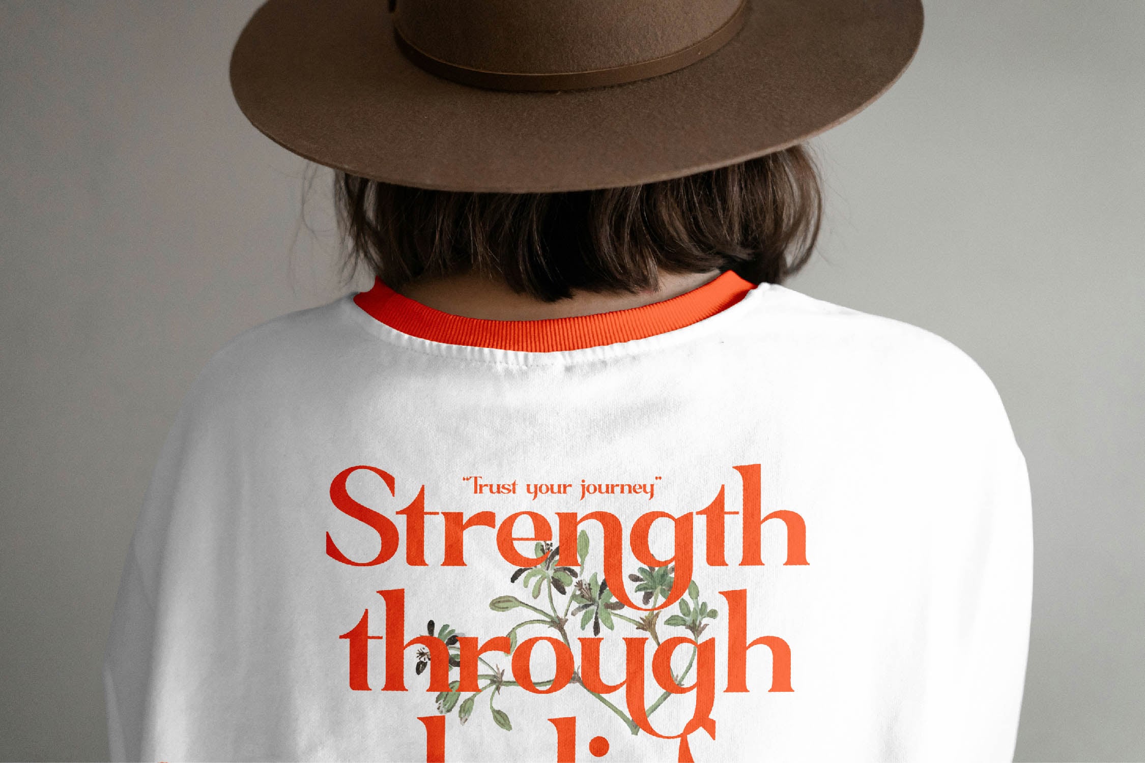 Back view of white tee with orange ringer collar, red serif text, botanical illustration, brown hat