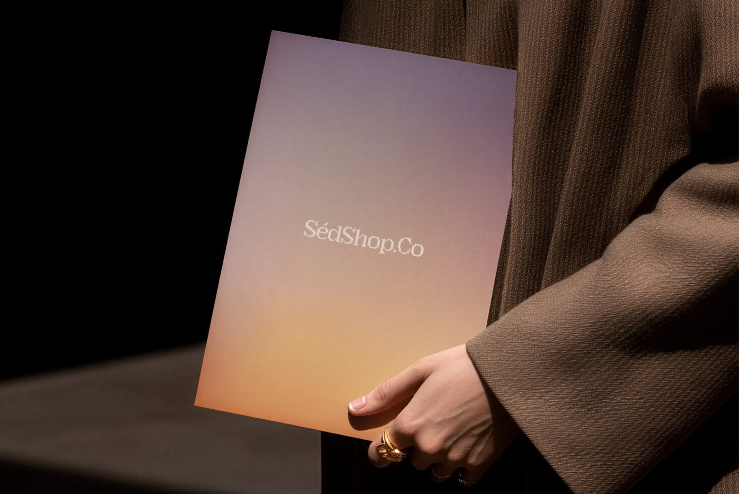Hand holding gradient brochure with SédShop.Co logo in warm peachy tones