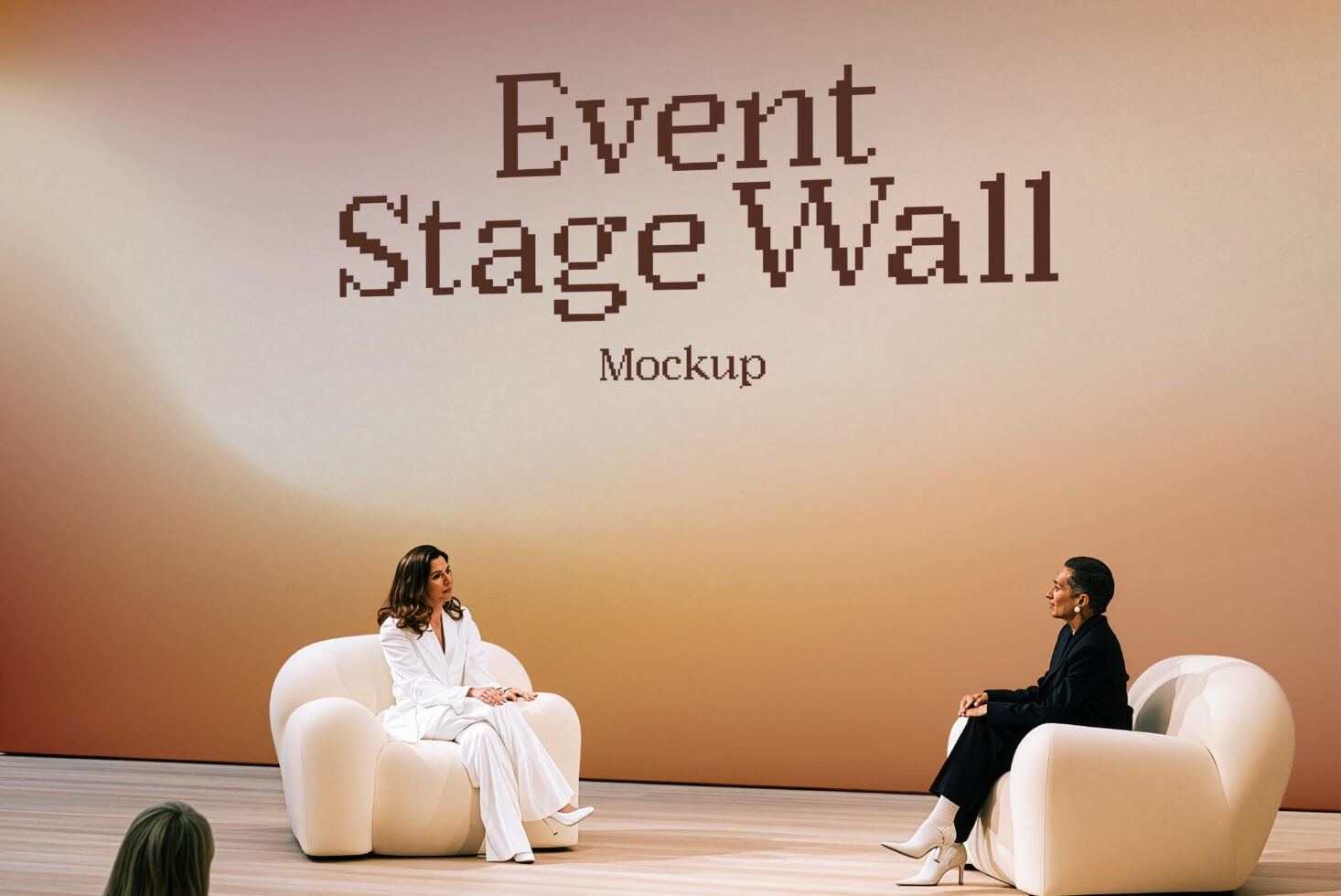 Two women chatting on cream chairs facing stage wall with pixelated Event Stage Wall text
