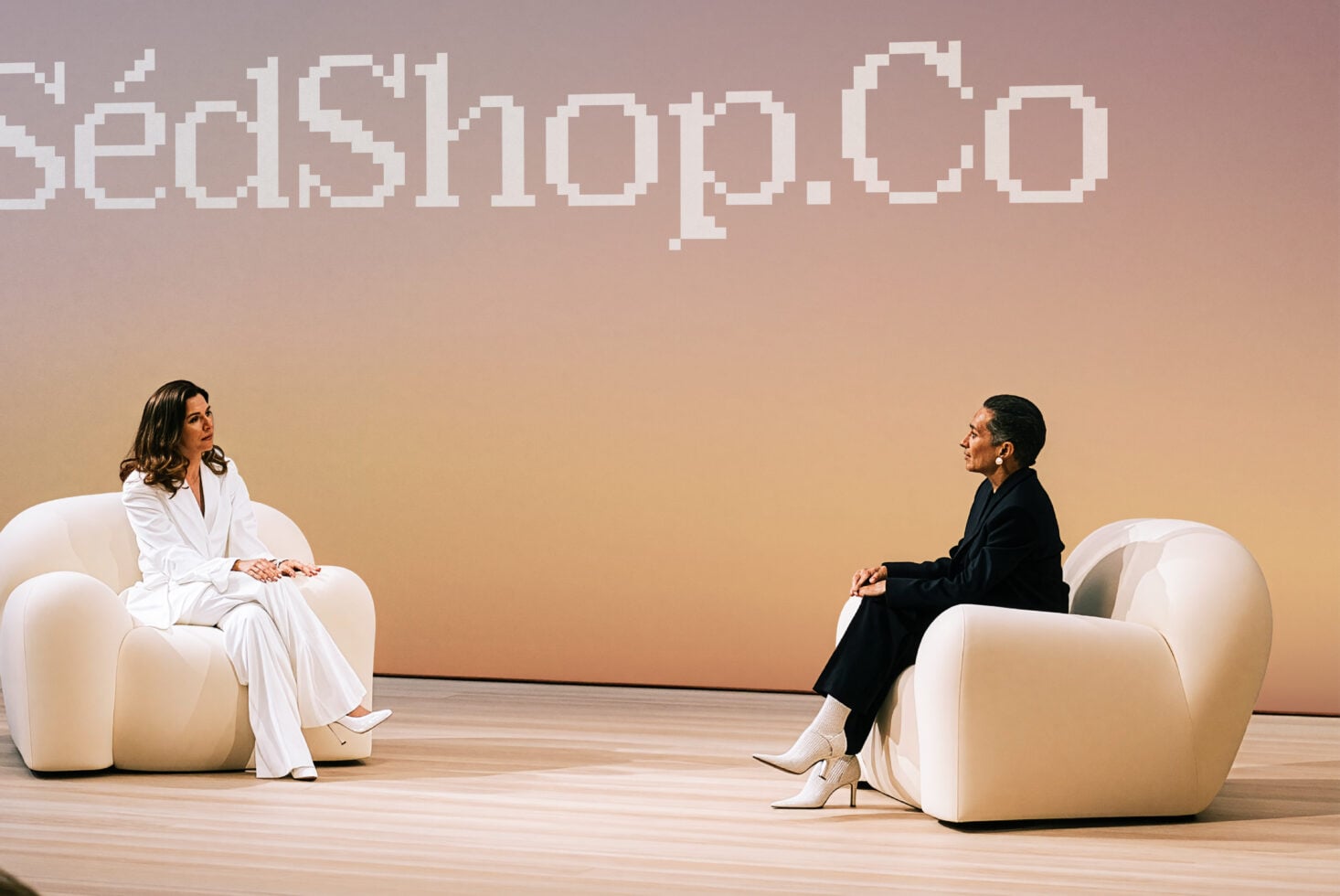 Two women sitting in curved cream chairs facing each other with pixel art text above on warm gradient background