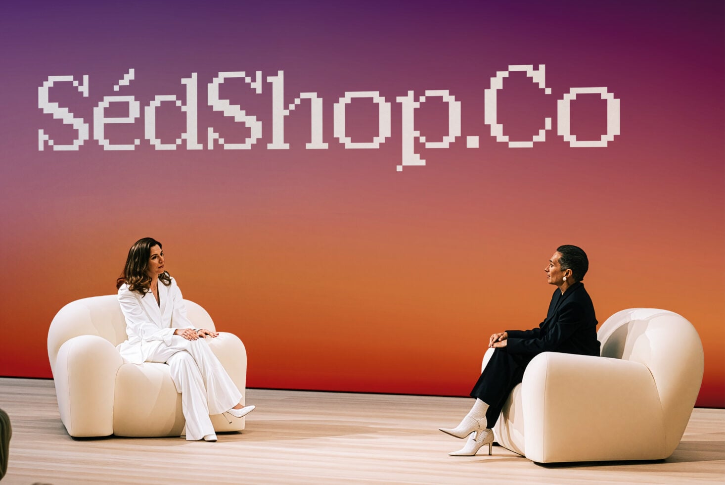 Two women sitting in cream chairs facing each other on stage with pixel art text glowing above them