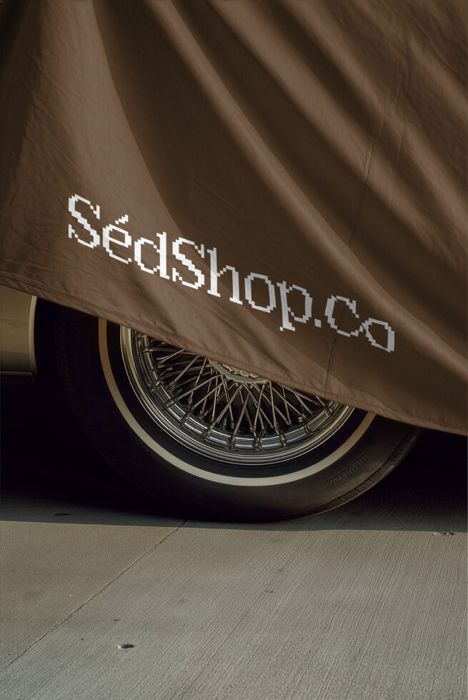 Beige car cover with pixelated branding text draped over chrome spoke wheel in sunlight