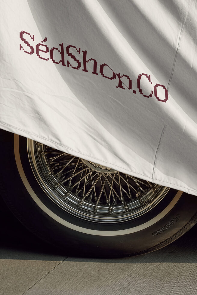 White car cover with maroon pixelated text draped over classic wire wheel in sunlight