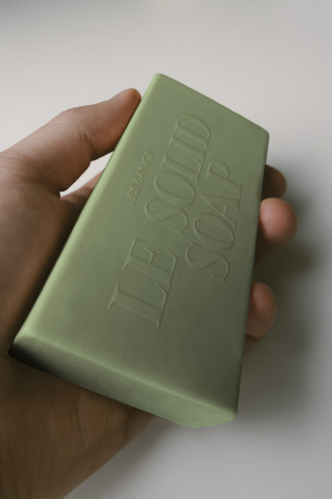Hand holding sage green soap bar with embossed logo text in realistic product shot