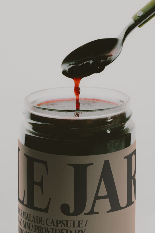 Paintbrush dripping red paint into open jar with beige label on white background