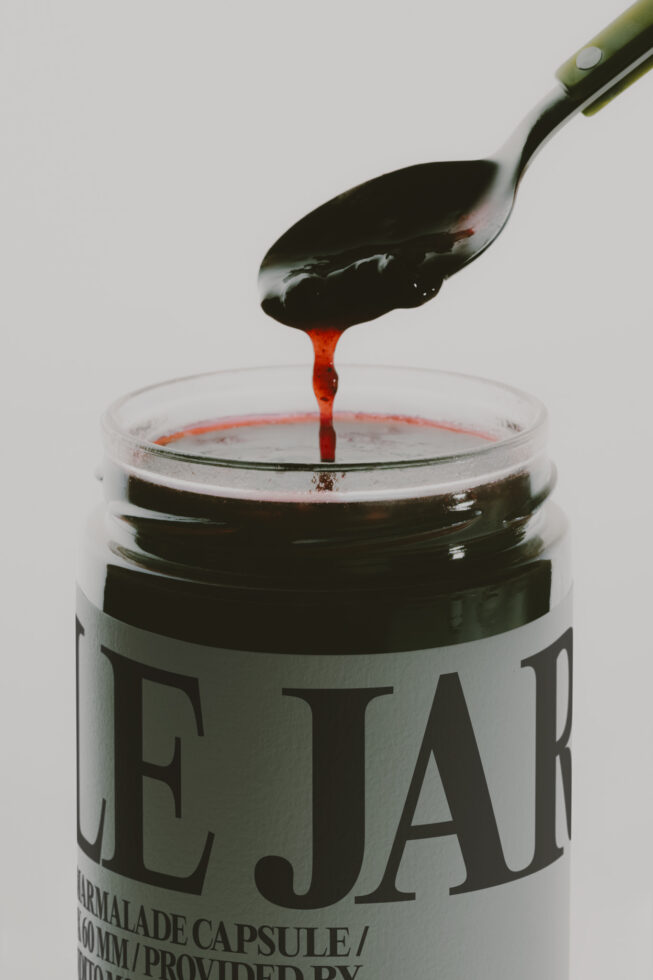 Paintbrush dripping red liquid into an open jar with black label on white background