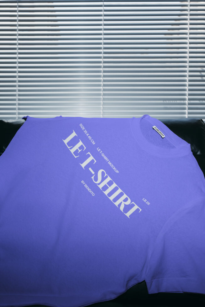 Bright purple tee with cream diagonal text draped on black couch, blinds behind