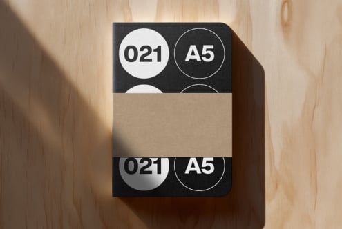 A5 notebook mockup with black cover featuring circular badge designs, kraft paper band wrapper on wood surface