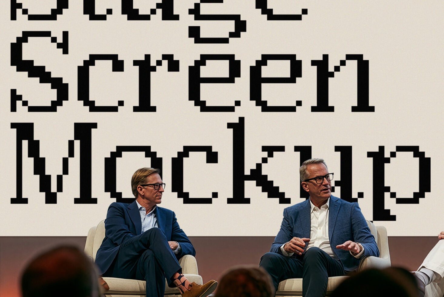 Two men in blazers sitting on beige couch with bold pixelated type on wall behind them