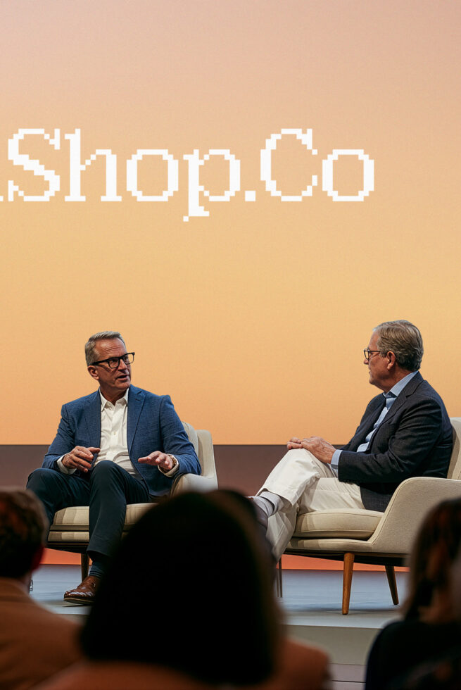 Two men in business casual sitting on stage chairs having a conversation with pixelated Shop.co text on warm beige wall behind them