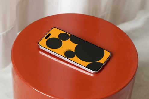 Smartphone mockup with bold yellow case displaying black circular camera cutouts, sitting on a rust-red cylindrical surface