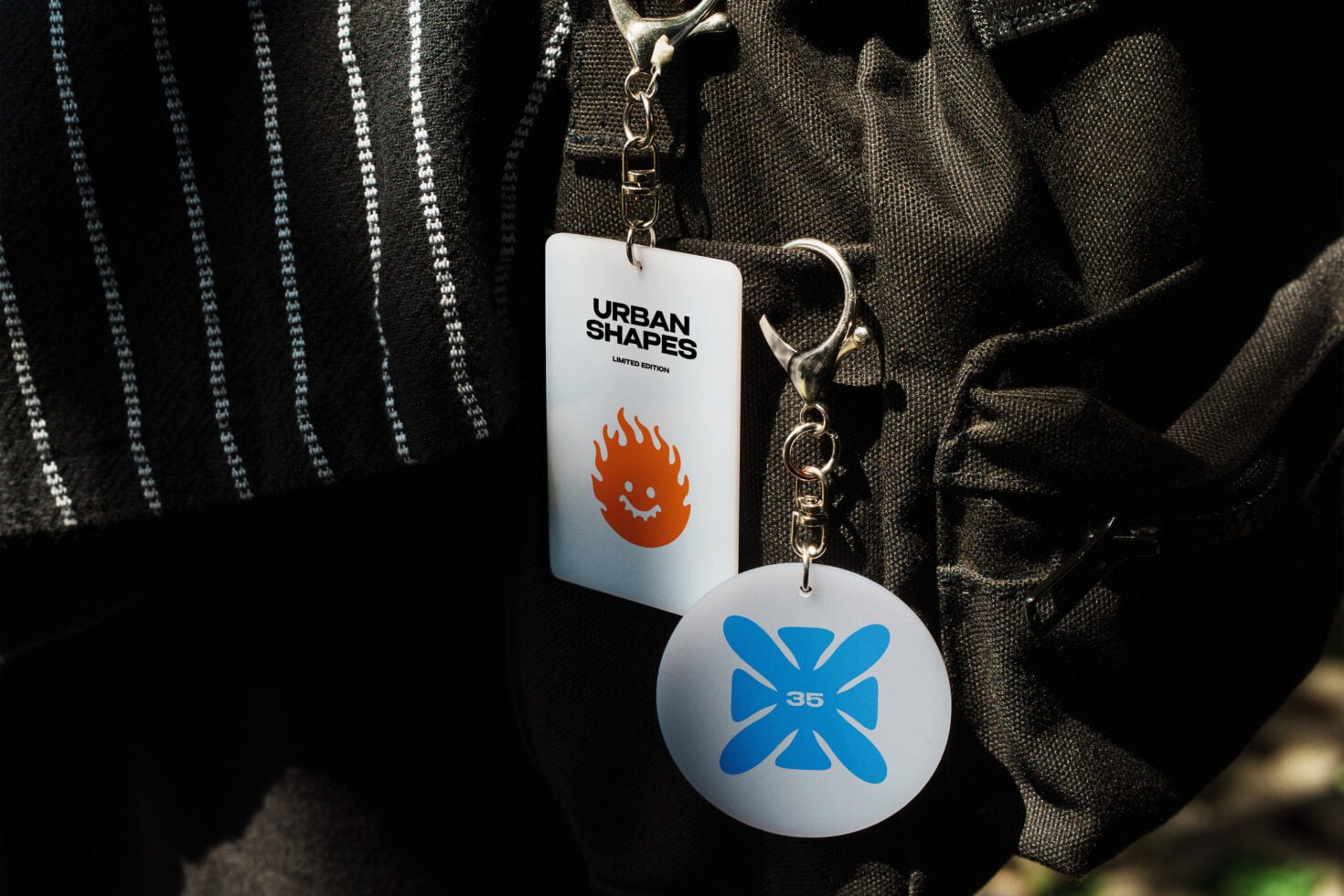 Bag charm keychain mockup with Urban Shapes branding, red flame character on tag, blue flower design on circular pendant, gold hardware on dark striped fabric