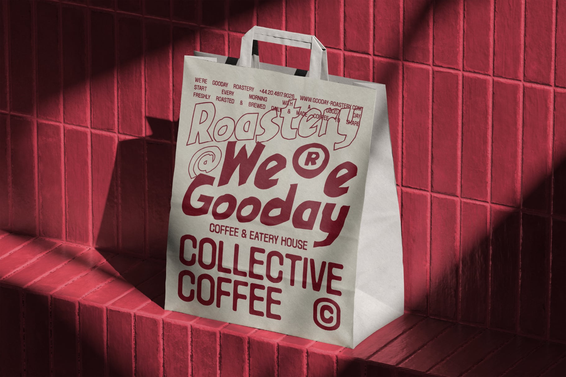 White paper tote bag with burgundy branding for Gooday Roastery coffee shop, sitting on red brick wall with dramatic shadows