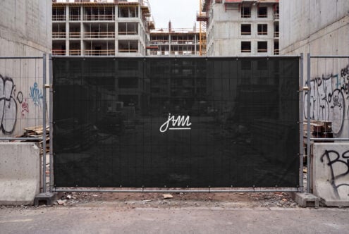 Black construction site gate with white cursive logo, concrete buildings in background