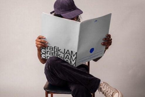 Person in purple corduroy outfit sitting on chair holding open magazine mockup with GrafikJAM branding visible