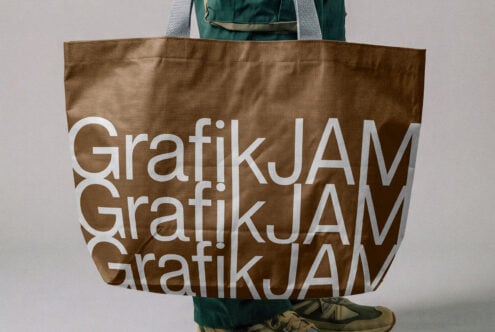 Tan canvas tote bag with white repeating Grafikjam text held by person in green jacket