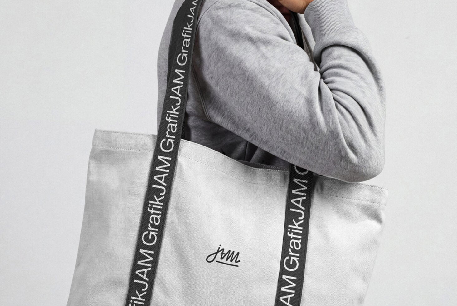Person wearing gray hoodie carrying white canvas tote bag with custom logo on branded strap