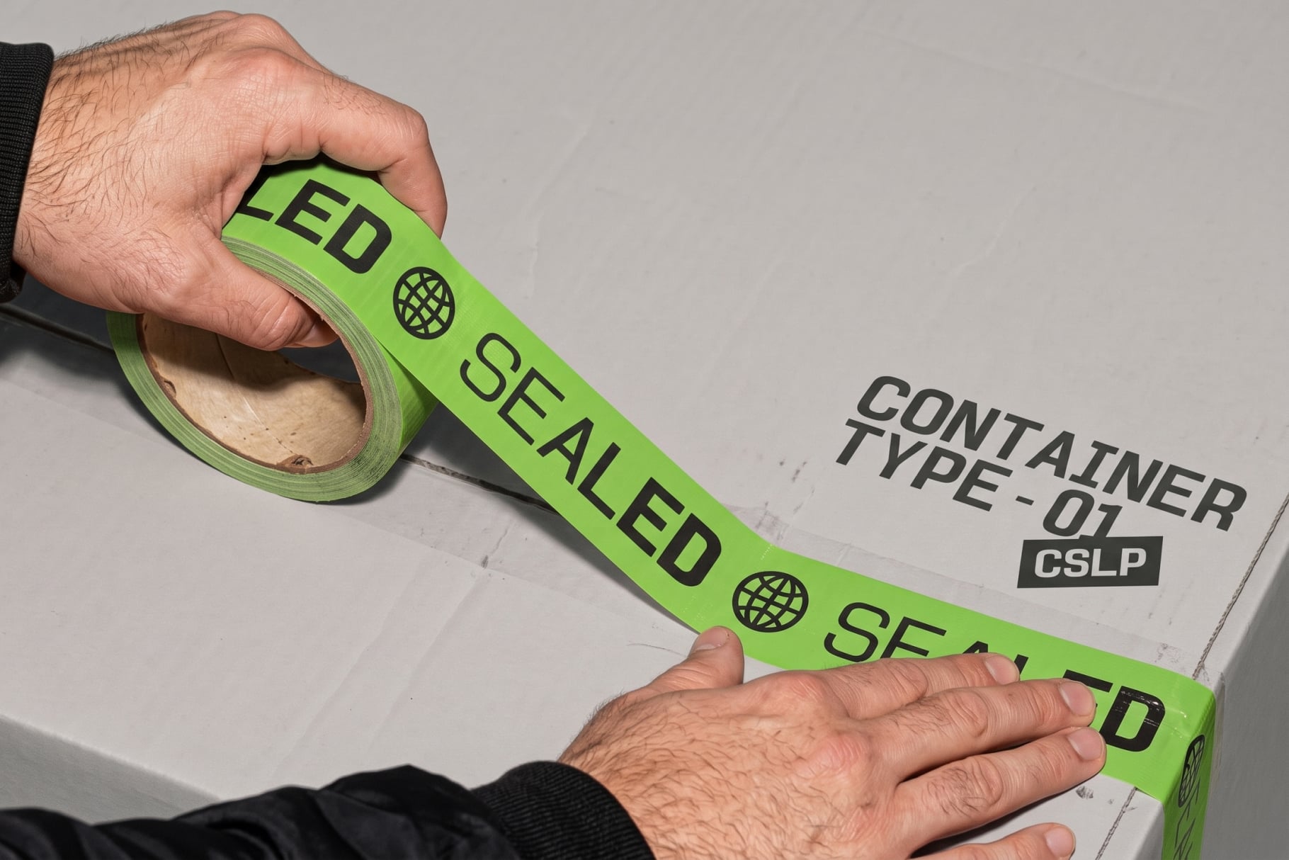 Hand unrolling bright green packing tape with black text across cardboard box mockup