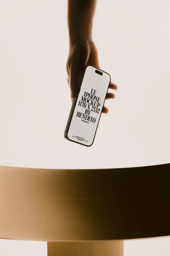 Hand holding iPhone 14 Pro Max mockup with white screen over minimal gold table setup