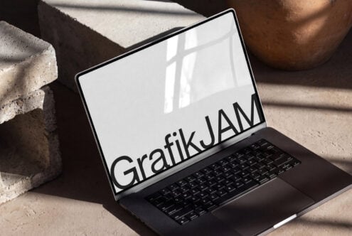 Laptop mockup with GrafikJAM text on screen, concrete blocks beside it, natural window light casting shadows