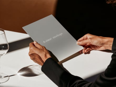 Hands holding gray notebook with "A vision mockups" text on white table with glass, warm lighting