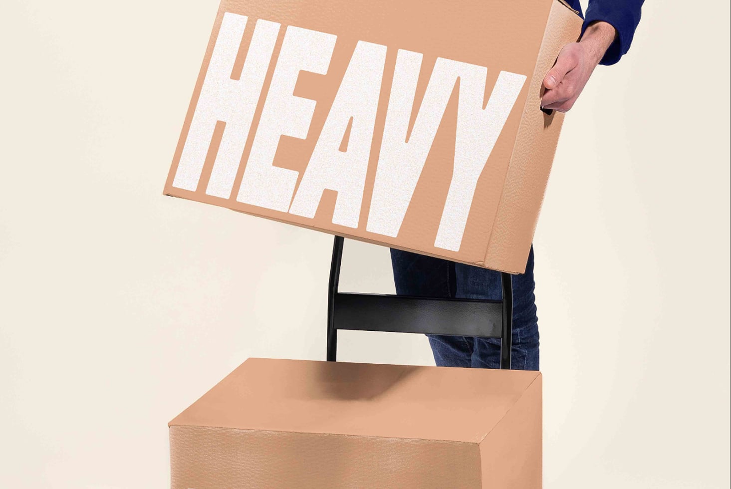 Person holding large tan cardboard box with bold white "HEAVY" text above wooden crate