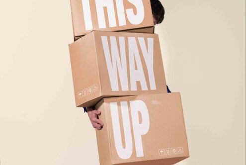 Person holding stacked kraft boxes with bold white letter cutouts spelling "THIS WAY UP"