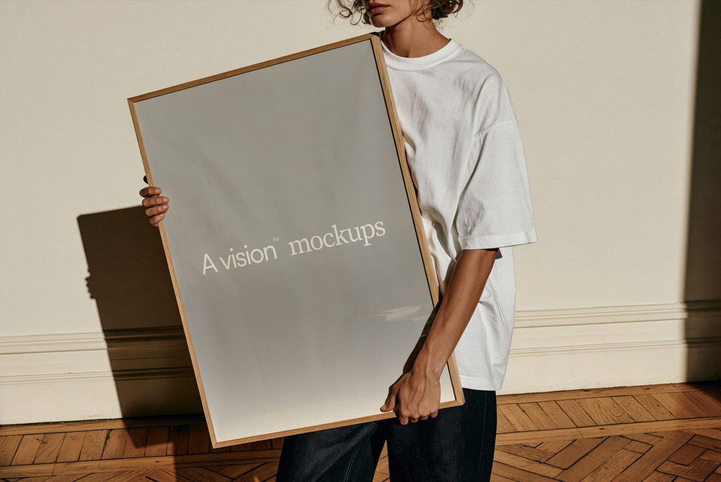 Person holding large framed poster mockup in white tee, natural wood frame, gallery wall setup