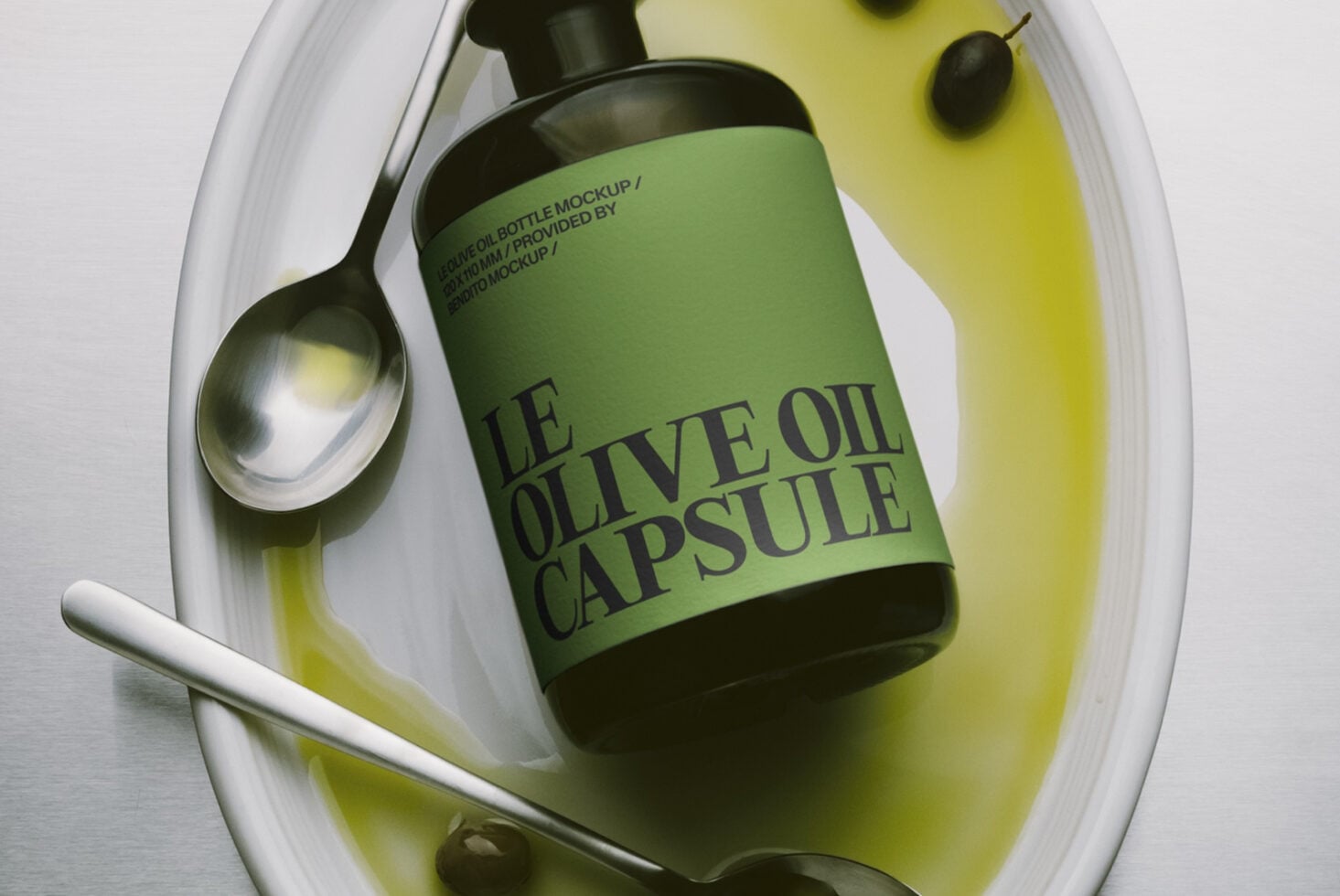 Olive oil capsule bottle mockup with green label sitting in white ceramic dish surrounded by spoon, pipette, oil puddles