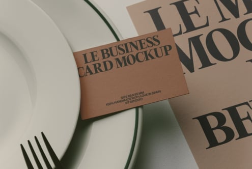 Tan business card mockup with serif typography, styled with magazines, fork, plus green plant stem on white surface