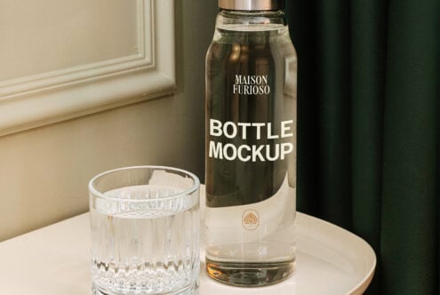 Luxury glass bottle with metal cap next to crystal tumbler on marble table, muted interior backdrop