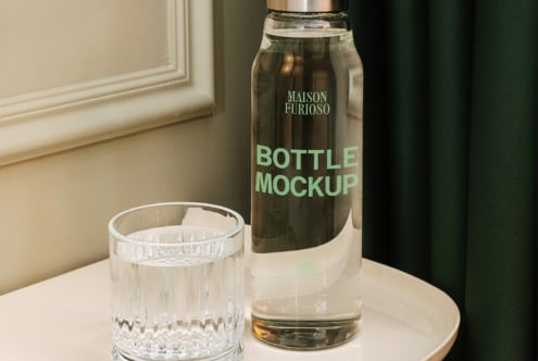 Glass bottle with mint label next to crystal tumbler on cream table, moody green backdrop