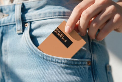 Hand holding tan business card with black label in jeans pocket, sunlit outdoor mockup