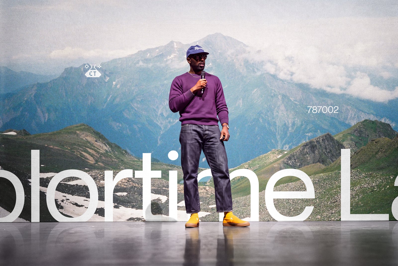 Guy speaking on stage with mountain backdrop, purple sweatshirt, yellow boots, bold white text overlay