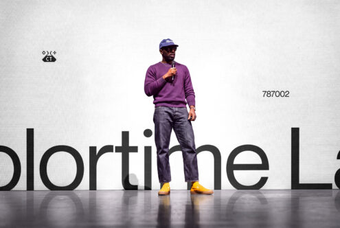 Man on stage holding microphone wearing purple sweatshirt, cap, with yellow shoes