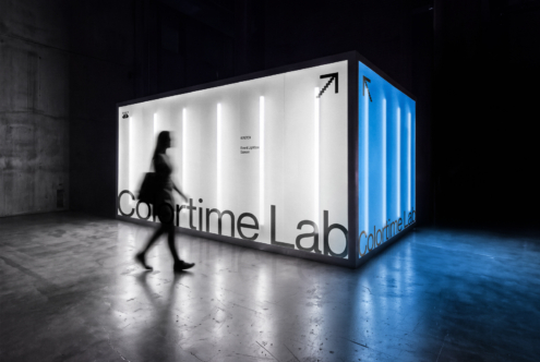 Illuminated exhibition booth with white panels, blue side, person walking past in dark warehouse space