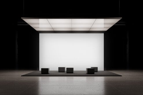 Minimalist trade show booth with blank white panel, overhead grid lighting, black seating cubes on platform