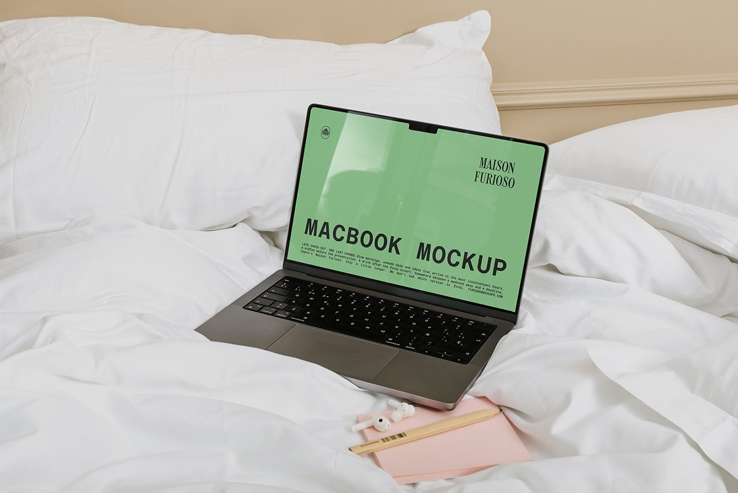 MacBook on white bedding with green screen mockup, pink notebook, pencils nearby