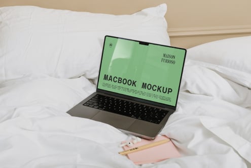 MacBook on white bedding with green screen mockup, pink notebook, pencils nearby